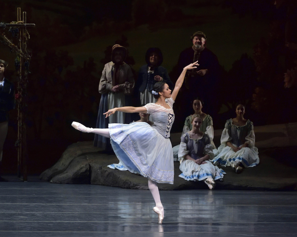 Misty Copeland dancing Giselle, wearing a soft blue Romantic tutu, Misty floats in an arabesque as the other dancers sit watching her in the background.